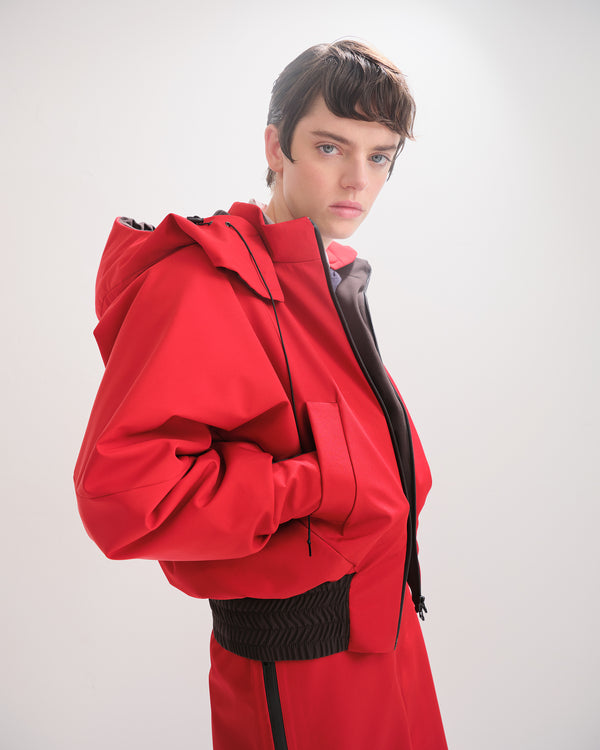 Person wearing a red  bomber jacket with detachable hood with black accents on the elasticated waist and cuffs on a white background