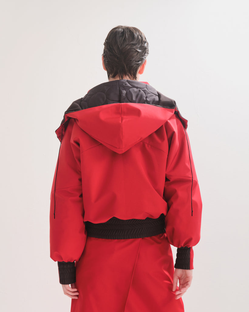 Back view of a person wearing a red  bomber jacket with detachable hood with black accents on the elasticated waist and cuffs on a white background