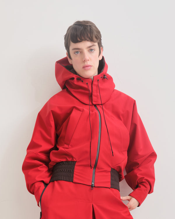 Person wearing a red  bomber jacket with detachable hood with adjustable cords and black accents on the elasticated waist and cuffs. 