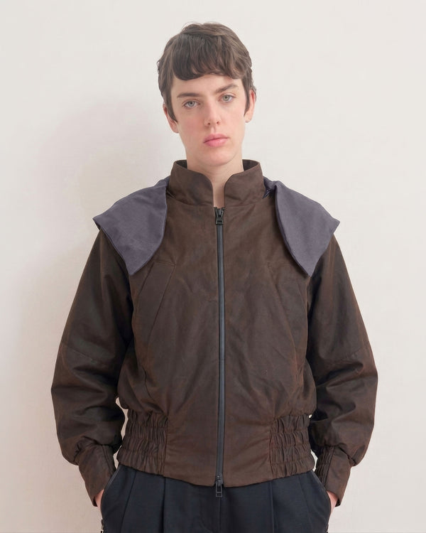 A model wearing a dark brown, waxed cotton jacket with contrasting dark gray shoulder panels that belong to the removable hood, and a black front zipper, paired with dark pants, set against a plain off-white background.