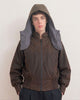A model wearing a dark brown, waxed cotton, hooded jacket with a unique integrated neck warmer collar in a contrasting gray fabric, set against a plain off-white background.
