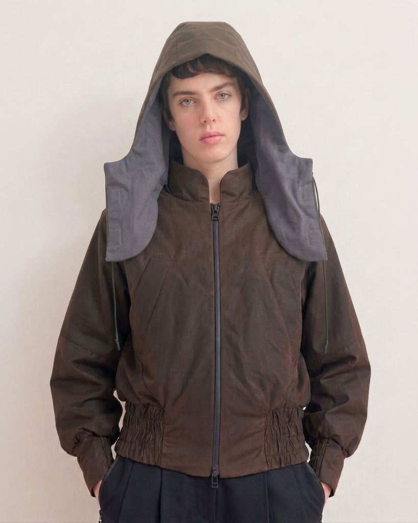A model wearing a dark brown, waxed cotton, hooded jacket with a unique integrated neck warmer collar in a contrasting gray fabric, set against a plain off-white background.