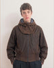 A model wearing a dark brown, waxed cotton, hooded jacket with a unique draped collar design, front zipper, and elasticized hem, paired with dark trousers against a plain light background.