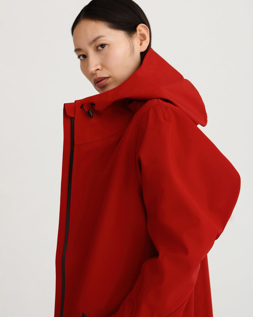 a side close up of midlength, red, waterproof, breathable, sustainable and technical raincoat from recycled materials with hood.